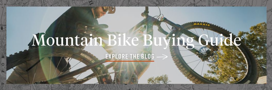 The sun shines through a mountain bike frame. The text reads: Mountain Bike Buying Guide. Explore The Blog